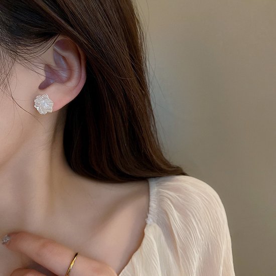 Een model draagt witte bloem-vormige oorknoppen met gedetailleerde blaadjes. Ze draagt een crème blouse en een gouden ring.