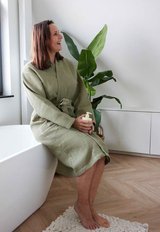 Vrouw in olijfgroene wafel badjas, zittend naast een badkuip met een flesje lotion.