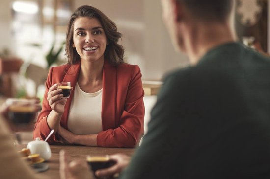 Een lachende vrouw in rode blazer met een glas espresso in haar hand aan tafel met twee mannen.