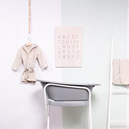 Beige badjas met stippenpatroon hangt aan een haakje. Naast een grijs babybadje met een alfabetposter.