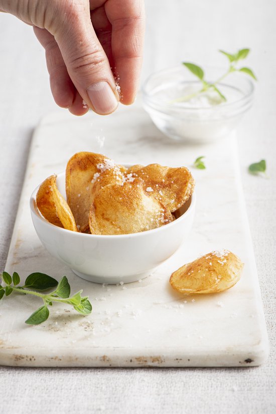 Hand strooit zout over gouden, krokante frietjes in een wit kommetje op een wit dienblad.