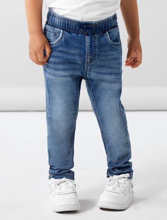 Jongen draagt een lichtblauwe spijkerbroek met trekkoord en witte sneakers.