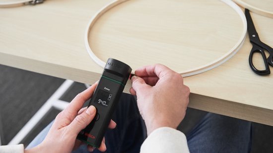 Handen gebruiken de Bosch Zamo afstandsmeter met lintadapter om de omtrek van een ronde vorm te meten.