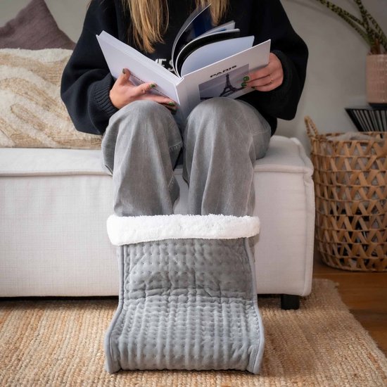 Grijze voetenwarmer met witte pluche voering, gedragen door een persoon zittend op een bank, lezend.