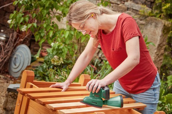 Vrouw schuurt een houten tuinmeubel met een groene Bosch multischuurmachine. Stofopvangbak bevestigd.