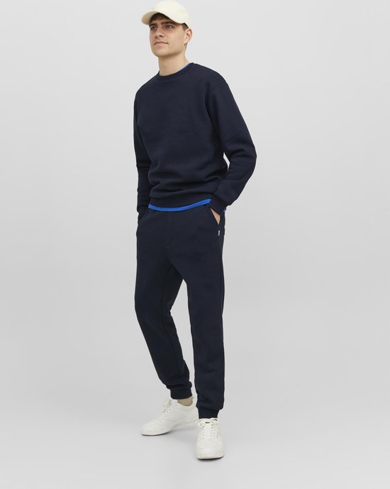 Model draagt een donkerblauwe sweater met ronde hals, met een blauwe rand zichtbaar en een witte pet.