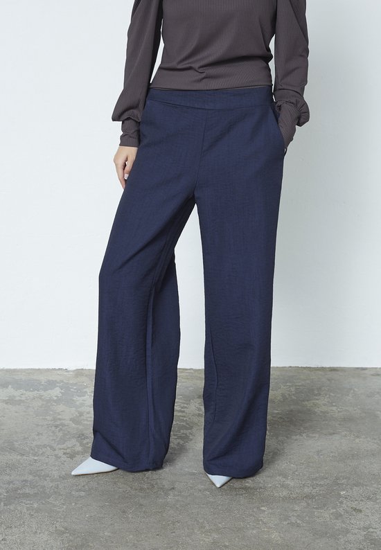 Model draagt een donkerblauwe broek met wijde pijpen en steekzakken.