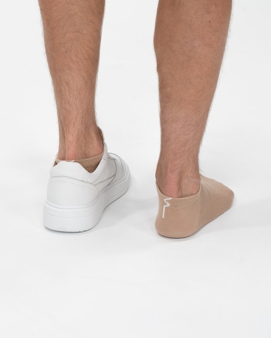 Blote benen met beige enkelsokken. Een voet draagt een witte sneaker.
