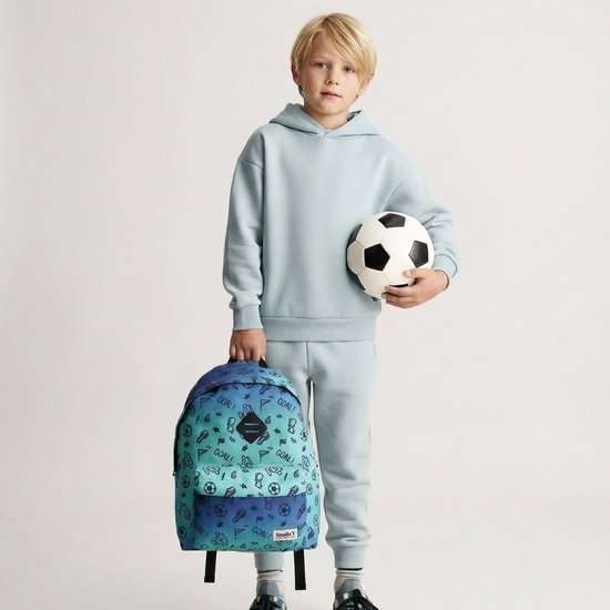 Jongen met blonde haren, gekleed in een lichtblauwe joggingpak, houdt een voetbal vast en draagt een blauw-groene rugzak met voetbalprint.