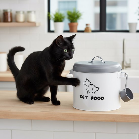 Zwarte kat met groene ogen kijkt naar een witte voercontainer met grijze deksel en schep.