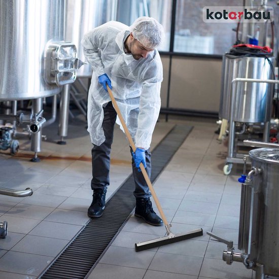 Een man in beschermende kleding gebruikt een vloertrekker met houten steel in een industriële omgeving.