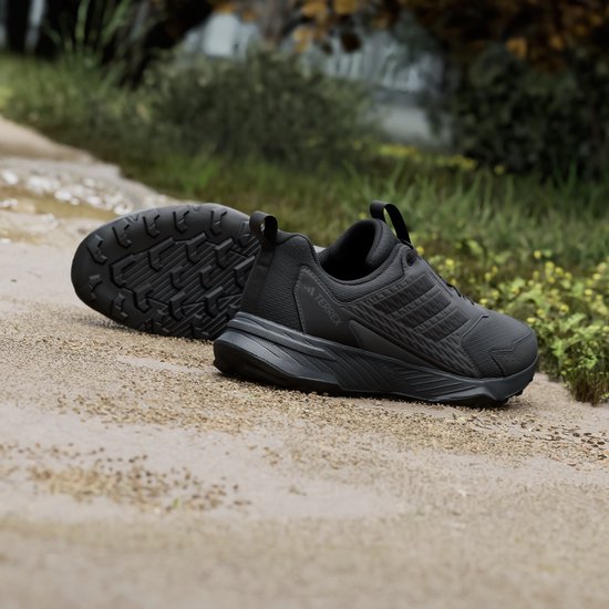 Paar zwarte Adidas Terrex trailrunningschoenen op zand, de zolen zichtbaar.