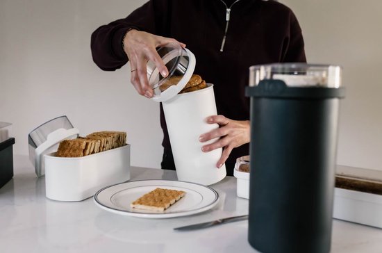 Een witte broodtrommel wordt gevuld met beschuit. Naast staat een zwarte bus met rijstwafels. Een persoon met een donkere trui vult de bus.