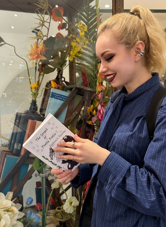 Een jonge vrouw met blond haar in een paardenstaart, draagt een blauw gestreept shirt, leest een boek met de titel 'The Gospel of Poetic Frolic'.