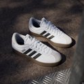 Witte Adidas VL Court 3.0 sneakers met zwarte strepen en bruine zool, gefotografeerd op een betonnen ondergrond.
