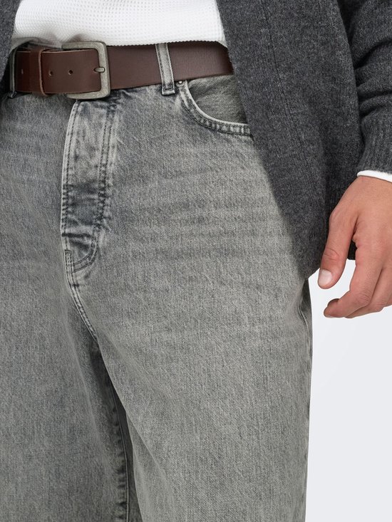 Detail van een grijze jeans met een bruine riem en een man die zijn hand in zijn zak steekt.