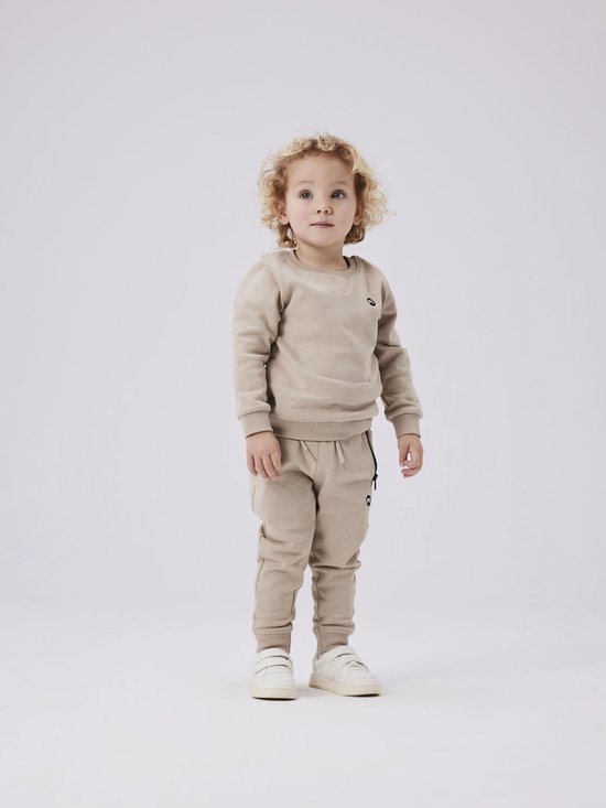 Jongen met krullend blond haar draagt een beige joggingbroek en sweater.