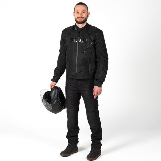 Man met zwarte motorjas met DXR-logo, jeans en helm in de hand.