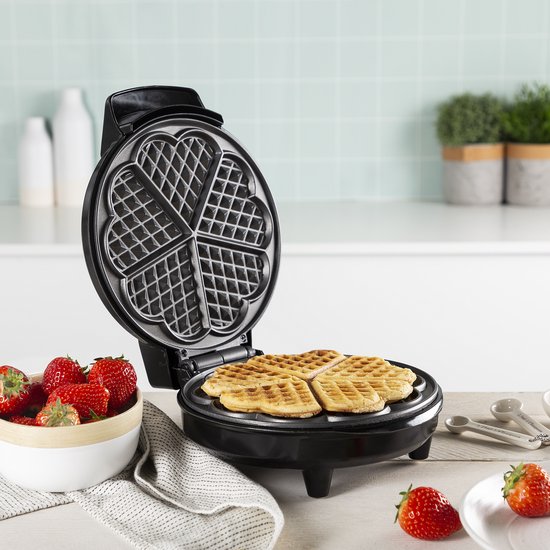 Open zwart wafelijzer met 4 hartvormige wafels. Een schaal met aardbeien staat ernaast.
