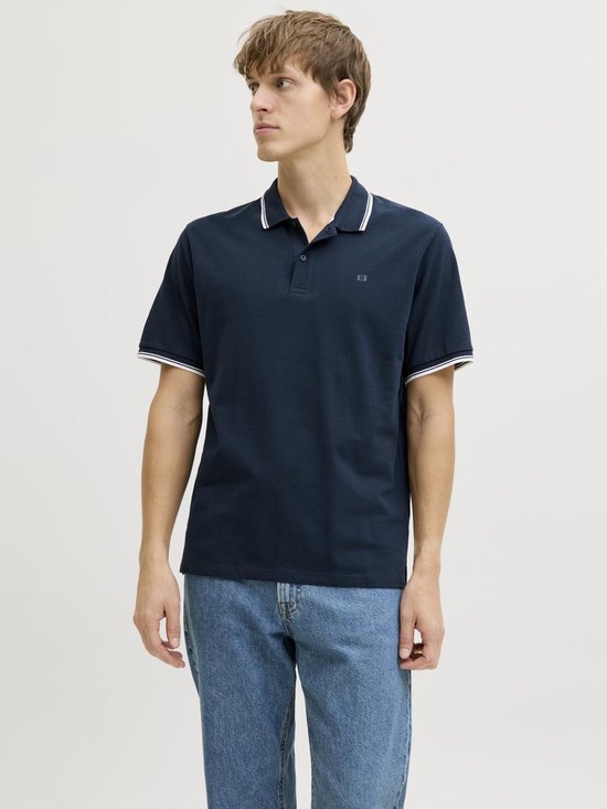 Man in donkerblauw poloshirt met witte streepdetails op kraag en mouwen, en blauwe jeans.