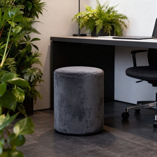 Donkergrijze fluwelen poef, rond, naast een bureau met planten.