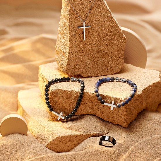 Drie sieraden op zand en stenen: een zilverkleurige ketting met kruis, een zwarte kralenarmband met kruis en een blauwe kralenarmband met kruis.
