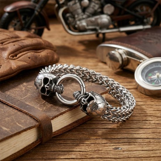 Zilverkleurige schakelarmband met dubbele schedel en ring sluiting op een houten ondergrond met een notitieboekje.