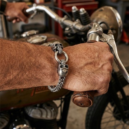 Zilverkleurige schakelarmband met schedelmotief, gedragen aan een arm naast een motorfiets.