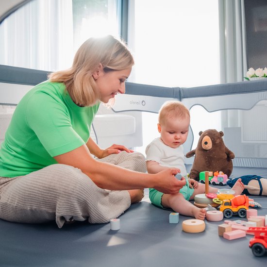 Vrouw met baby in grijze box, speelt met speelgoed. De box heeft een berenmotief.