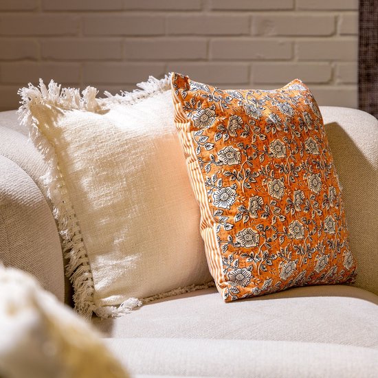Sierkussen met oranje bloemenprint en witte streepjes, naast een crème kussen met franjes.