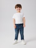 Jongetje met kort bruin haar draagt een wit T-shirt en een donkerblauwe spijkerbroek met stonewashed effect, trekkoord en omgeslagen broekspijpen.