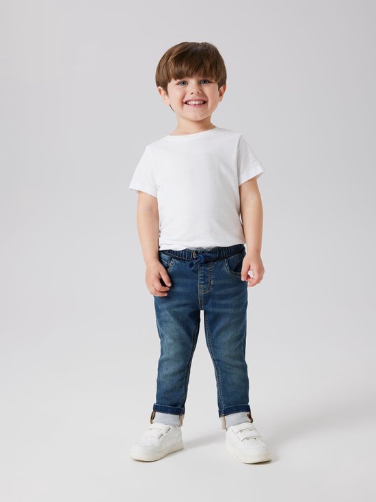 Jongetje met kort bruin haar draagt een wit T-shirt en een donkerblauwe spijkerbroek met stonewashed effect, trekkoord en omgeslagen broekspijpen.