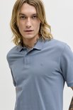 Close-up van een man met blond haar die een blauwgrijze polo draagt, met zichtbare tatoeages op nek en arm.