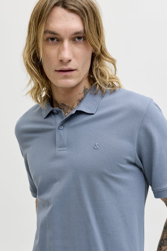 Close-up van een man met blond haar die een blauwgrijze polo draagt, met zichtbare tatoeages op nek en arm.