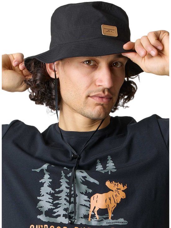 Man met zwarte vissershoed en T-shirt met elandprint. Hij pakt de rand van de hoed vast.