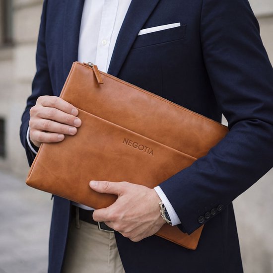 Man met donkerblauw jasje en beige broek draagt cognac leren laptoptas met 'NEGOTIA' logo.