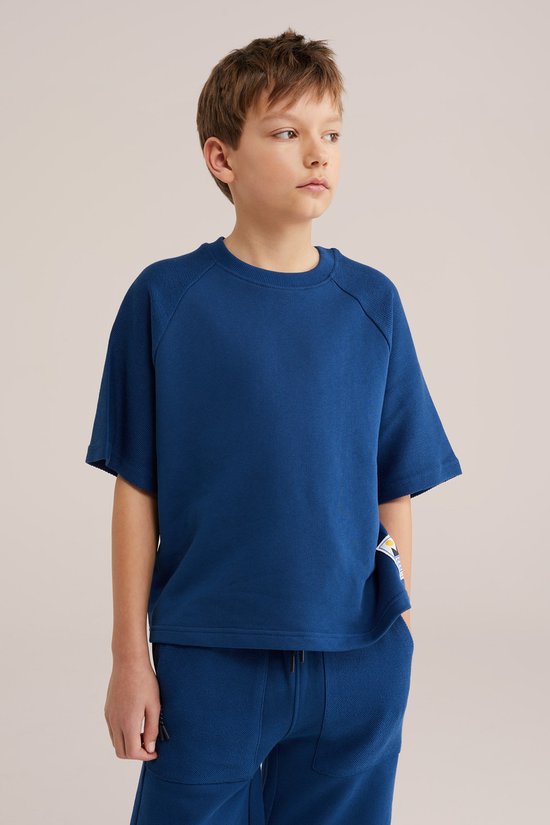 Jongen draagt een kobaltblauwe sweater met korte mouwen en een ronde hals. Hij draagt ook een bijpassende kobaltblauwe korte broek.