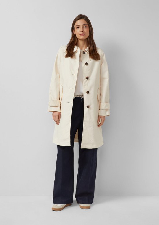 Vrouw in crèmekleurige s.Oliver trenchcoat, wit T-shirt en navy broek.