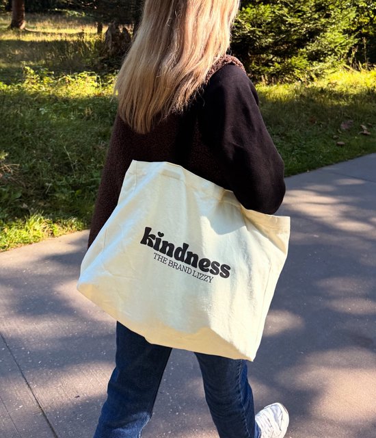 Sac shopper en toile écru avec texte noir 'kindness THE BRAND LIZZY'. Porté sur l'épaule par une fille aux cheveux blonds, portant une veste marron et un jean.