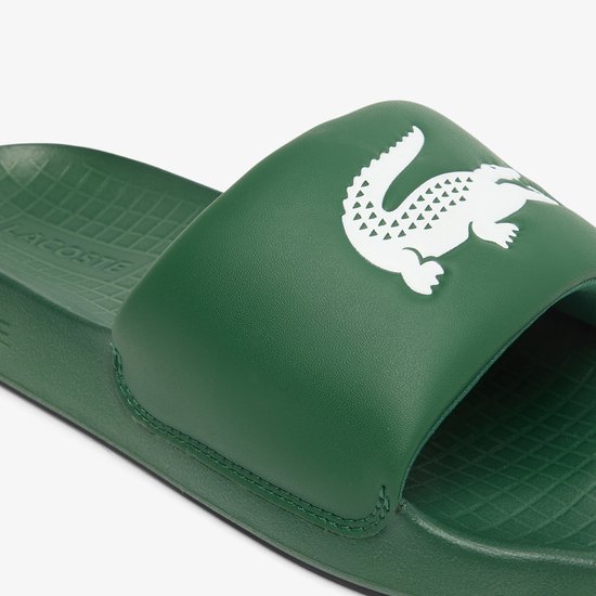 Close-up van een donkergroene Lacoste slide met een prominent wit krokodillenlogo op de band. De binnenzool heeft een rasterpatroon.