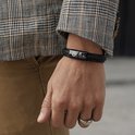 Zwarte gevlochten leren armband met zwart gespsluiting, gedragen door een man met een geruite blazer.
