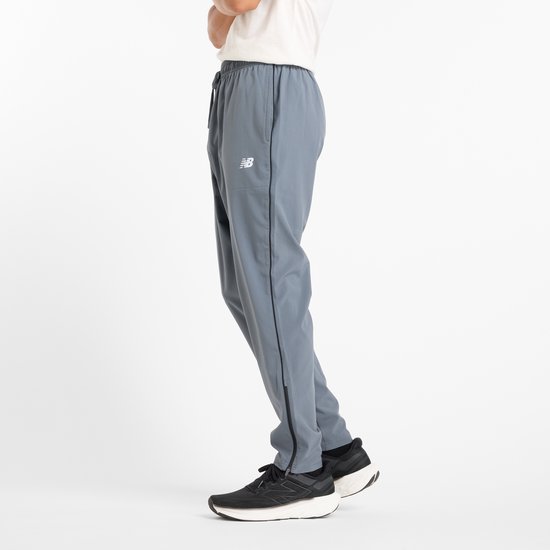 Grijze New Balance sportbroek met zwarte accenten, gedragen door een model met witte t-shirt en zwarte sneakers.