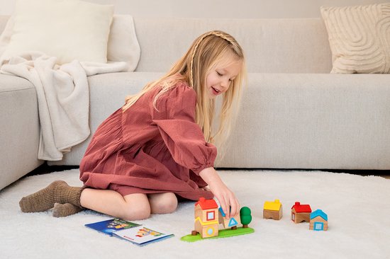 Meisje speelt met houten blokken: rode, gele en blauwe huizen, op een witte vloer, met een opdrachtenboekje.