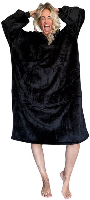 Een vrouw met blonde haren trekt aan haar zwarte, oversized hoodie deken, die tot op de kuiten valt. Ze staat op blote voeten.