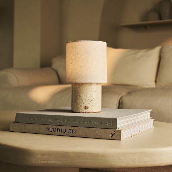 Tafellamp met beige linnenkap en cilindrische travertijnen voet, staand op stapel boeken.