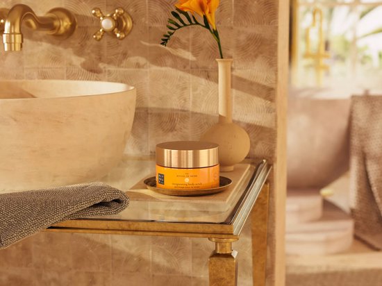 Een oranje pot 'The Ritual of Mehr' body scrub staat op een bijzettafel naast een handdoek, een wastafel en een vaas met een bloem.
