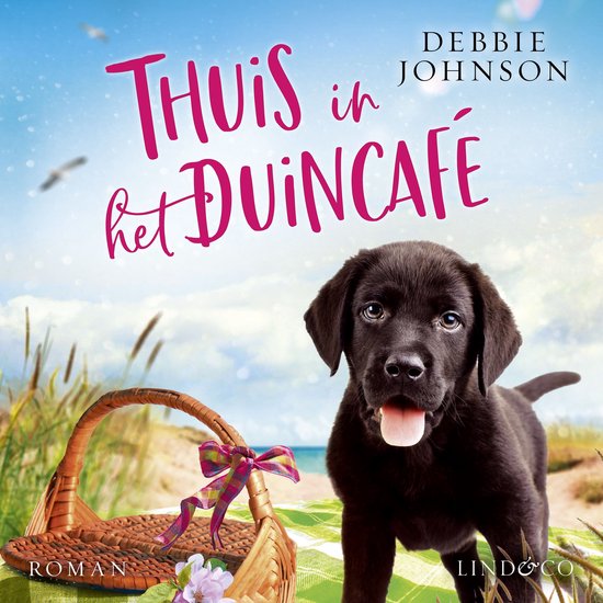 Zwarte labrador puppy naast een picknickmand met strik, op een deken aan zee. Titel: Thuis in het Duincafé.