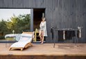 Zwart droogrek met kleding op een houten terras bij een modern huis. Vrouw staat bij de deur.