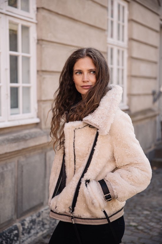Femme aux longues boucles brunes, porte une veste en shearling crème avec des sangles et des boucles en cuir noir.