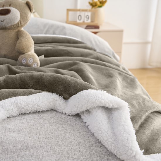 Taupe fleece plaid met witte sherpavoring, met een teddybeer en decoratieve kalender op de achtergrond.